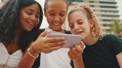 Fototapeta premium Three girls friends pre-teenage are sitting on the waterfront using mobile phone, watching videos, photos. Three teenagers on the outdoors in urban cityscape background