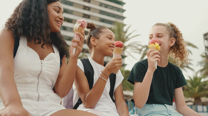 Laughing three girls friends pre-teenage are sitting on the waterfront testing ice cream. Three...
