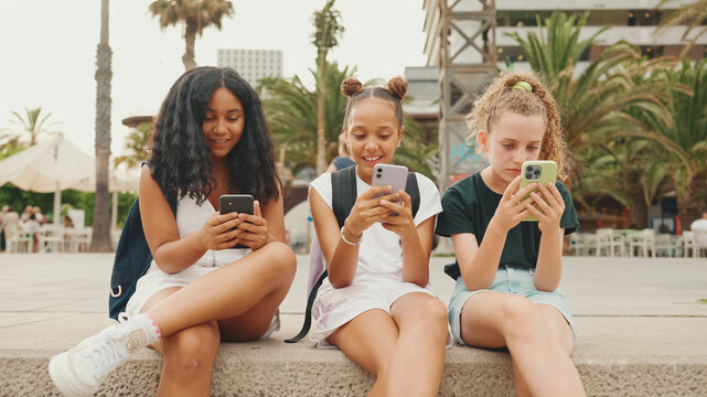 Three girls friends pre-teenage are sitting on the waterfront using mobile phone. Three teenagers playing games on a smartphone on the outdoors in urban cityscape background - Powered by Adobe