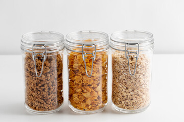 food storage, healthy eating and diet concept - jars with oat, corn flakes and granola on white...