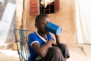 African village boy sitting in a sunny courtyard drinking from a big blue plastic mug; scarcity of water supply to private households in West African landlocked countries