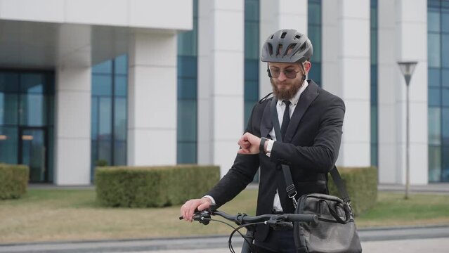 A handsome man in a suit looks at his watch, standing near the business center. Man wearing a bicycle helmet and shoulder bag, standing near black old-fashioned bicycle. High quality 4k footage