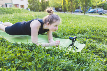 Young fit blonde woman practicing plank on yoga mat and recording video tutorial on camera outdoor on sunny summer day. Fit gen z girl doing yoga on grass in park. Healthy lifestyle, online yoga