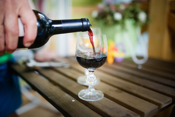 Hand with bottle of wine pours red wine into glass on wooden table on terrace in summer