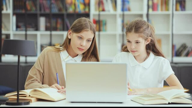 School children wearing uniform sitting at desk, looking at laptop and writing in exercise books, doing assignment in class together, bookshelves on blurred background. Concept of education