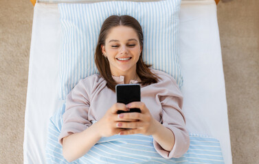 people, bedtime and rest concept - happy smiling teenage girl with smartphone lying in bed at home