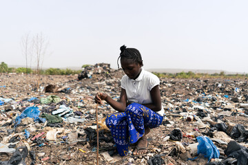 African girl sitting in the middle of heaps of waste, in charge of finding recyclable materials such as metal, glass and plastic; informal waste recycling; exploitation - obrazy, fototapety, plakaty