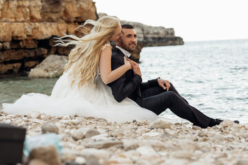 Side view of beautiful couple sitting on stones pebbles at shore near water. Young woman bride hugging kissing groom.