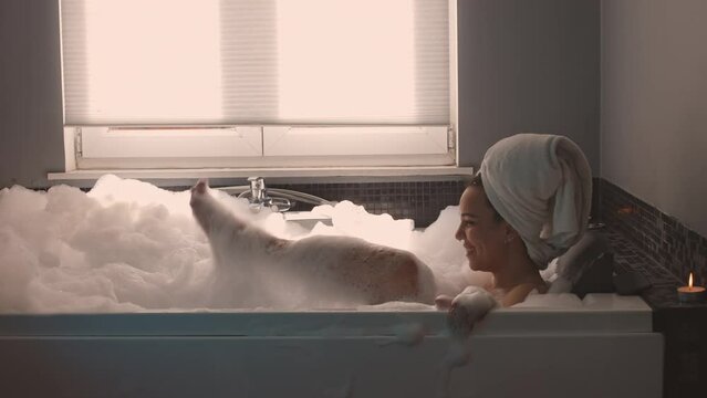 young Brazilian lady in a cinematic shot style relaxing in a bath filled with bubbles - girl in relaxing warm a foam bathtub at hotel - concept of leisure, rest, comfort, skincare, healthcare