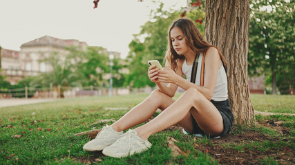 Naklejka na ściany i meble Beautiful girl with long wavy hair wearing in white top uses mobile phone while sitting on the green grass under tree in the park. Girl flip through the pictures on your phone with fingers