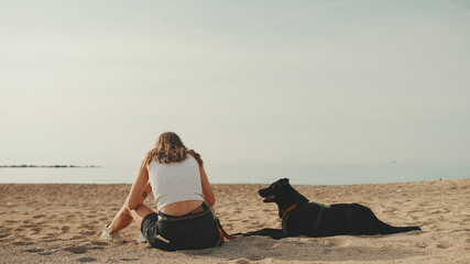 Cute girl with long wavy hair in white top plays on pebble beach with black dog seascape background
