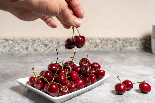 detail of a hand picking a cherry from a plate full of fresh red cherries on a stone kitchen worktop. Cherry seasonal fruit