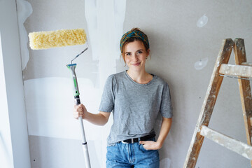 Thinking young woman renovates her apartment and has a painter role in hand, looking at camera