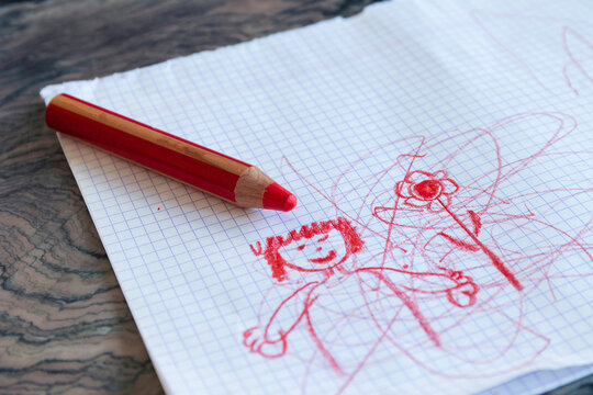 A portrait of a large thick red coloring pencil for small children lying on a white checkered sheet or page with a cute toddler drawing or doodle on it.
