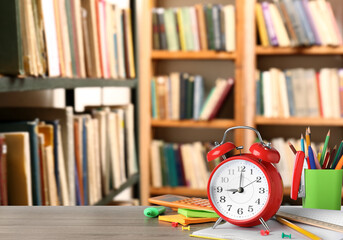 Red alarm clock and different stationery on wooden table in library, space for text
