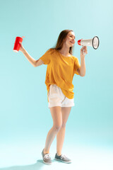 Full-length portrait of young beautiful girl in casual cloth, shouting in megaphone, posing isolated over blue studio background