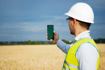 an engineer is working on his smartphone in the field, an agricultural engineer is standing in the field, an engineer is holding a phone in his hand, an engineer is wearing a helmet