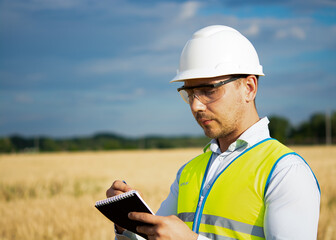 Fototapeta premium engineer in a helmet, glasses and a yellow vest, hand on a helmet, holding a notebook, a folder, making notes in a notebook