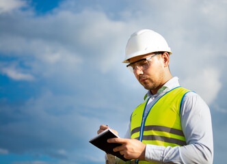 Fototapeta premium engineer in a helmet, glasses and a yellow vest, hand on a helmet, holding a notebook, a folder, making notes in a notebook