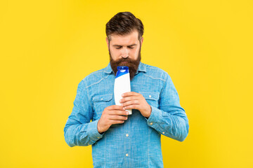 brutal bearded man smelling shampoo bottle on yellow background, presenting toiletries