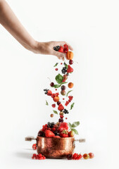 Woman hand pouring various summer berries into copper cooking pot: strawberries, currants, raspberries, cherries, gooseberries and apricots at white background.  Jam preparation