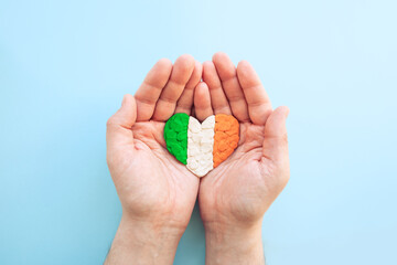 Flag of Ireland. Heart shape of green, white and orange plasticine modeling clay in male hands on blue background. Irish flag concept, love. Top view, closeup, copy space.