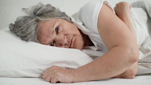 Senior Woman Without Emotions Lying In Bed Suffering From Insomnia. Close-up Of An Older Woman Relaxing In Bedroom, Looking At Camera And Around.