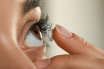 Young woman putting contact lens in her eye on grey background, closeup