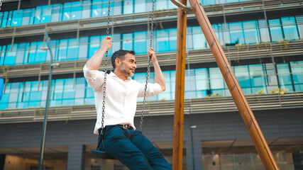 mature businessman with neat beard wearing white shirt swings and rides on children's swing. Successful man resting after hard day's work