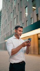 Mature businessman with neat beard uses mobile phone sits on bench in the financial district in the city. Successful man scrolls through information on smartphone and smiles