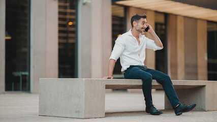 Mature businessman with neat beard wearing white shirt uses mobile phone sits on bench in the financial district in the city. Successful man talking on smartphone.