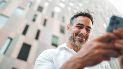 Mature businessman with neat beard uses mobile phone sits on bench in the financial district in the city. Successful man scrolls through information on smartphone and smiles