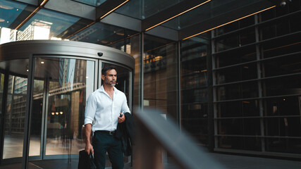 Fototapeta premium Mature businessman with neat beard wearing white shirt leaves the office in the financial district in the city. Successful man with mobile phone in his hands, after hard day's work