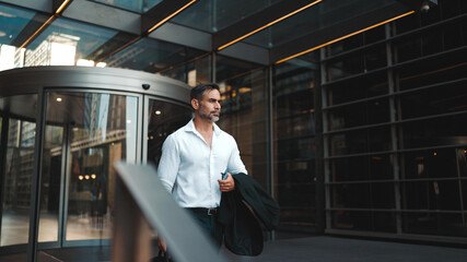 Mature businessman with neat beard wearing white shirt leaves the office in the financial district in the city. Successful man with mobile phone in his hands, after hard day's work