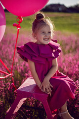 beautiful girl child of 5 years old is sitting on a pink stepladder in a pink dress in a field with...