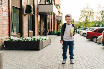 Back to school. Funny child with backpack holding notepad and training books going to school. Boy pupil with bag. Elementary school student going to classes. Kid walking outdoor on the city street.