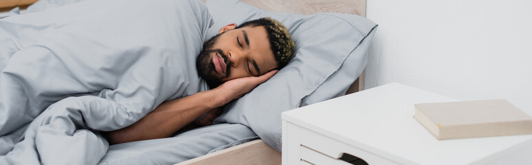 Fototapeta na wymiar bearded african american man with closed eyes sleeping in bed near bedside table with book, banner.