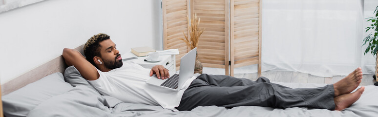 african american man in wireless earphone watching film while lying on bed with laptop, banner.