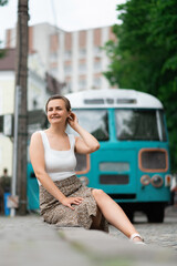 Fototapeta na wymiar An attractive young woman in a white blouse and brown skirt is sitting on a sidewalk in the background of an old school bus