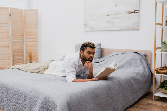 young african american man with dyed hair reading book while lying on bed.