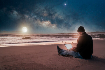 person sitting with laptop on the beach outdoors working under the starry night and milky way..