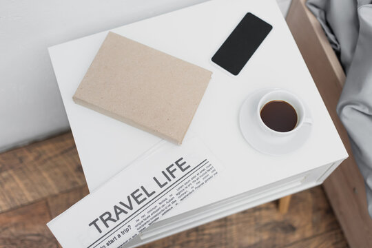 top view of cup with coffee and smartphone near newspaper with travel life lettering and book on white bedside table.