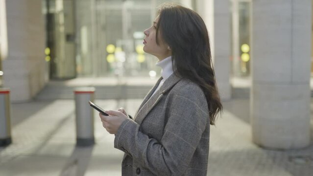 Camera Move Around: a young woman with dark hair stands and looks into the phone and around. Against the backdrop of office buildings. Slow motion 4k footage