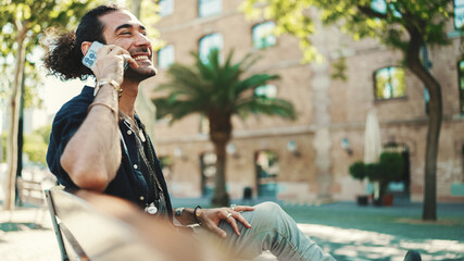 Attractive italian guy with ponytail and stubble is using mobile phone sitting on bench at old...