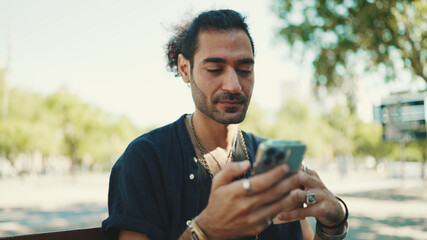 Attractive italian guy with ponytail and stubble is using mobile phone sitting on bench at old...