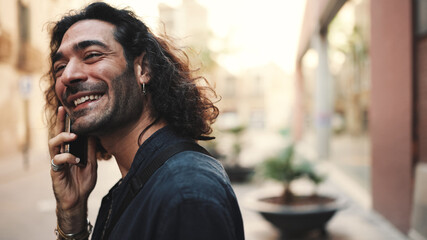 Young attractive italian guy with long curly hair and stubble is using mobile phone at old buildings background. Stylish man with an earring in his ear and lot of chains emotionally talking on phone