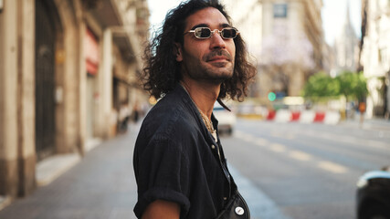 Young italian guy with long curly hair and stubble walks smiling down the street. Stylish man with...