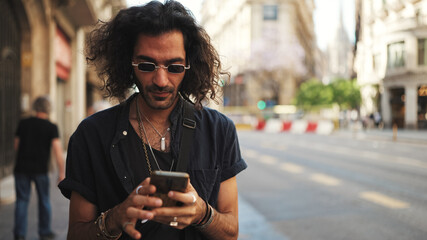 Young italian guy with long curly hair and stubble using mobile phone. Stylish man with sunglasses...