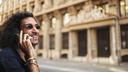 Fototapeta premium Young attractive italian guy with long curly hair and stubble is using mobile phone. Stylish man with talking on smartphone on busy street