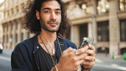 Young attractive italian guy with long curly hair and stubble is using mobile phone. Stylish man...